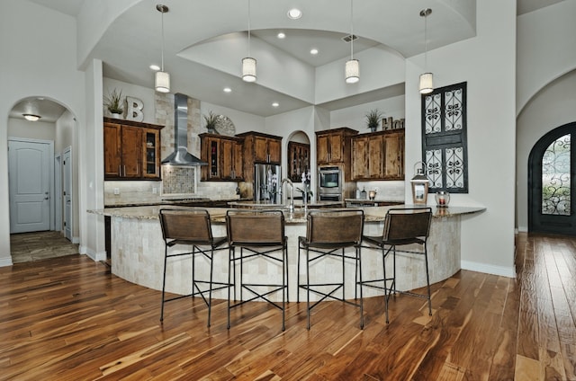 kitchen featuring appliances with stainless steel finishes, light stone counters, wall chimney range hood, dark wood-type flooring, and a towering ceiling