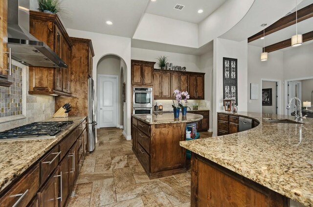 kitchen featuring wall chimney range hood, backsplash, hanging light fixtures, light stone counters, and a center island