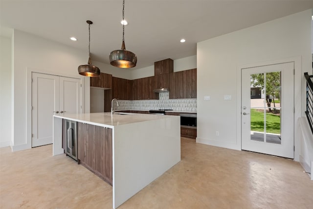 kitchen featuring beverage cooler, sink, decorative backsplash, dark brown cabinetry, and a center island with sink