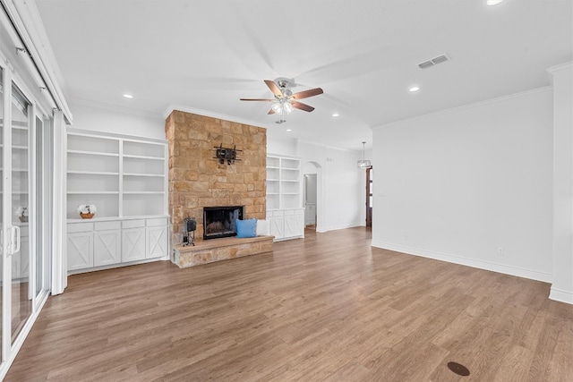 unfurnished living room featuring ceiling fan, built in shelves, ornamental molding, light hardwood / wood-style floors, and a stone fireplace