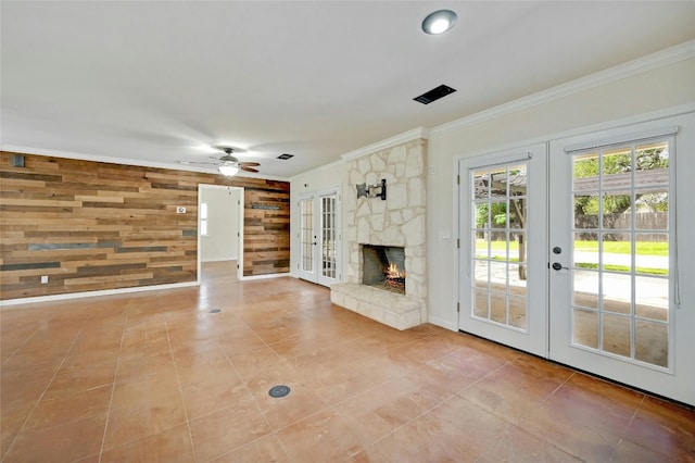 unfurnished living room with tile patterned flooring, french doors, wooden walls, ceiling fan, and a stone fireplace