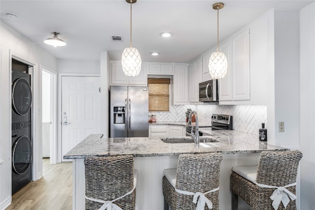 kitchen with stacked washing maching and dryer, appliances with stainless steel finishes, decorative light fixtures, sink, and white cabinets