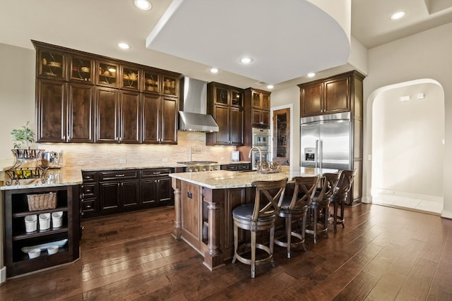 kitchen with appliances with stainless steel finishes, a center island with sink, wall chimney range hood, dark brown cabinetry, and dark hardwood / wood-style flooring