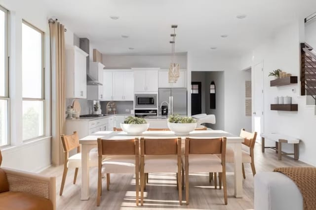 kitchen with appliances with stainless steel finishes, white cabinets, wall chimney range hood, light wood-type flooring, and an island with sink