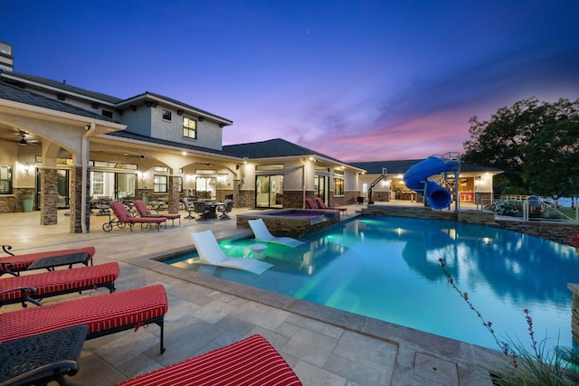 pool at dusk featuring an in ground hot tub, a patio area, a water slide, and ceiling fan