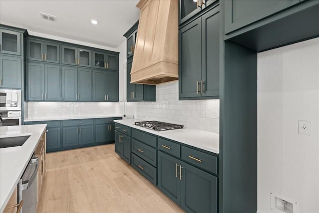 kitchen with stainless steel appliances, tasteful backsplash, light stone counters, light hardwood / wood-style floors, and custom exhaust hood