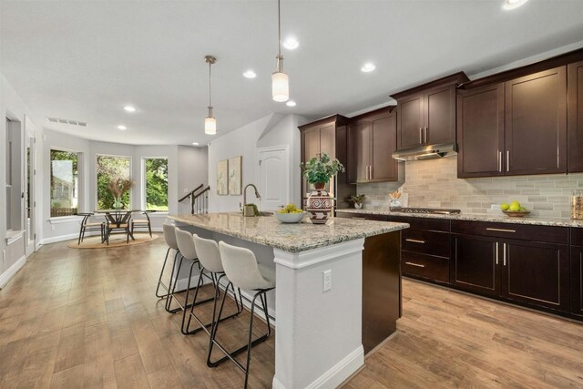 kitchen with light stone countertops, light wood-type flooring, pendant lighting, a center island with sink, and a breakfast bar area