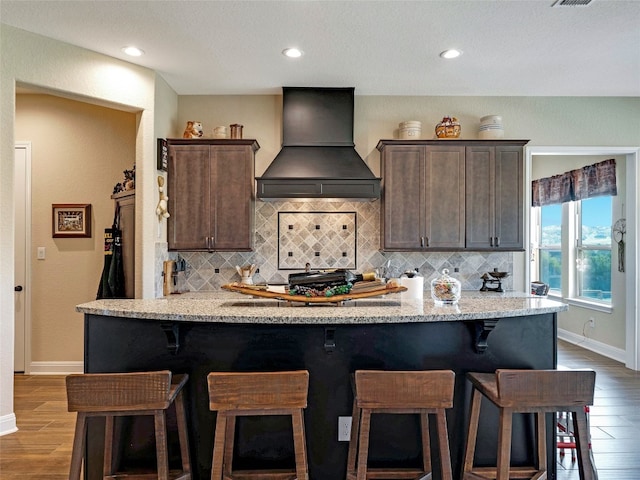 kitchen with dark brown cabinets, custom exhaust hood, hardwood / wood-style flooring, and light stone countertops