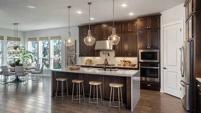 kitchen featuring dark wood-type flooring, hanging light fixtures, ventilation hood, a center island with sink, and appliances with stainless steel finishes