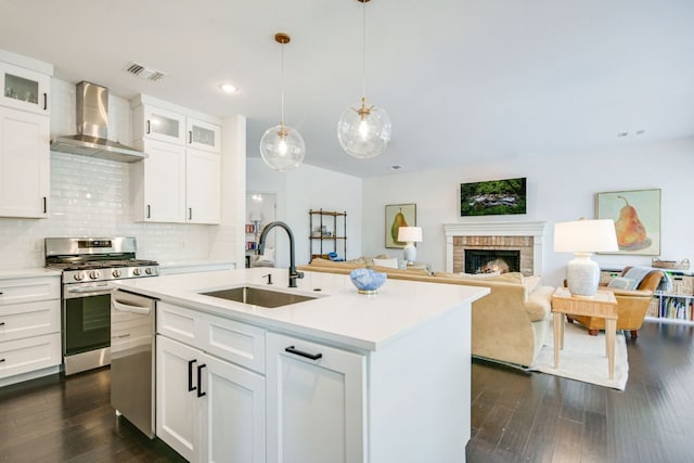 kitchen with sink, appliances with stainless steel finishes, an island with sink, white cabinets, and wall chimney exhaust hood