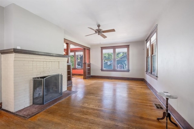 unfurnished living room with ceiling fan, wood finished floors, visible vents, baseboards, and a brick fireplace