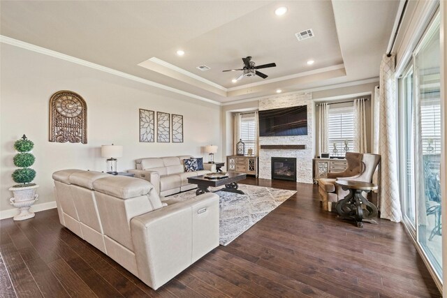 living room with a tray ceiling, a stone fireplace, dark wood-type flooring, ceiling fan, and ornamental molding