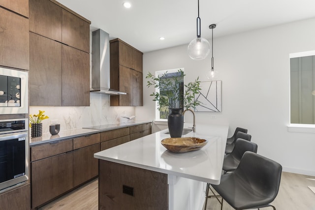 kitchen featuring black electric stovetop, stainless steel oven, light wood-type flooring, wall chimney exhaust hood, and a breakfast bar