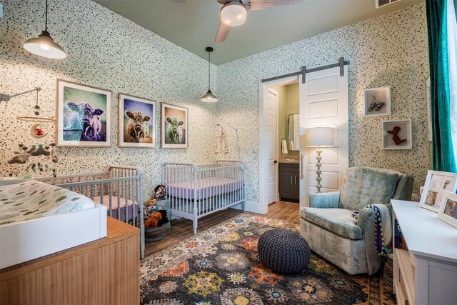 bedroom featuring a nursery area, hardwood / wood-style floors, ceiling fan, a barn door, and connected bathroom