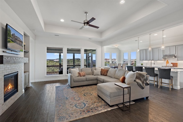 living room featuring a brick fireplace, a tray ceiling, dark hardwood / wood-style flooring, and ceiling fan