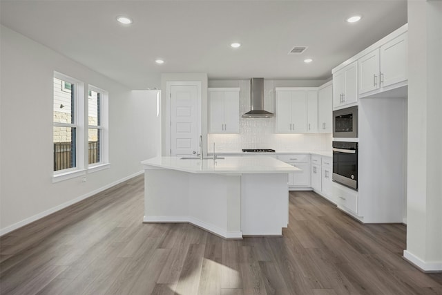 kitchen with stainless steel microwave, white cabinets, wall oven, a center island with sink, and wall chimney exhaust hood