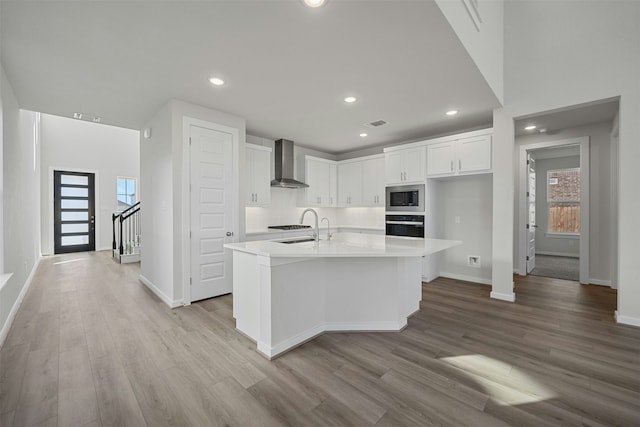 kitchen featuring wall chimney range hood, stainless steel microwave, an island with sink, white cabinets, and oven