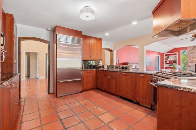 kitchen featuring sink, premium appliances, island range hood, kitchen peninsula, and dark stone counters