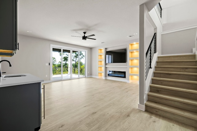 unfurnished living room with ceiling fan, sink, light wood-type flooring, and built in shelves