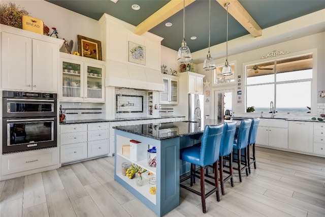 kitchen featuring decorative backsplash, beam ceiling, double oven, hanging light fixtures, and a center island with sink