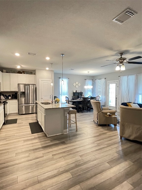 kitchen featuring ceiling fan with notable chandelier, an island with sink, white cabinets, light hardwood / wood-style floors, and stainless steel fridge