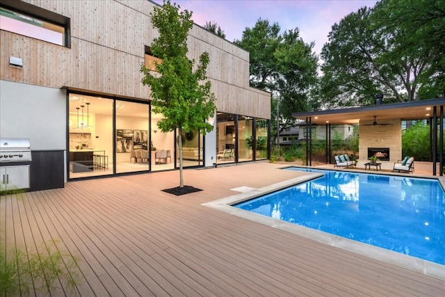 pool at dusk with ceiling fan, a wooden deck, grilling area, and exterior fireplace