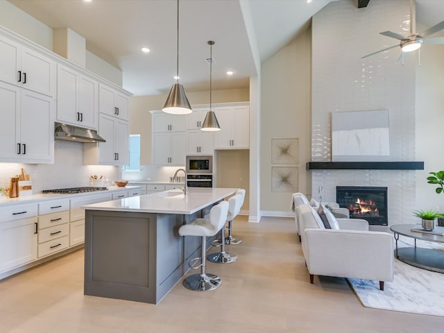 kitchen featuring a large fireplace, ceiling fan, white cabinetry, a center island with sink, and stainless steel appliances