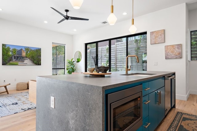 kitchen featuring stainless steel microwave, light wood-type flooring, a kitchen island with sink, hanging light fixtures, and sink