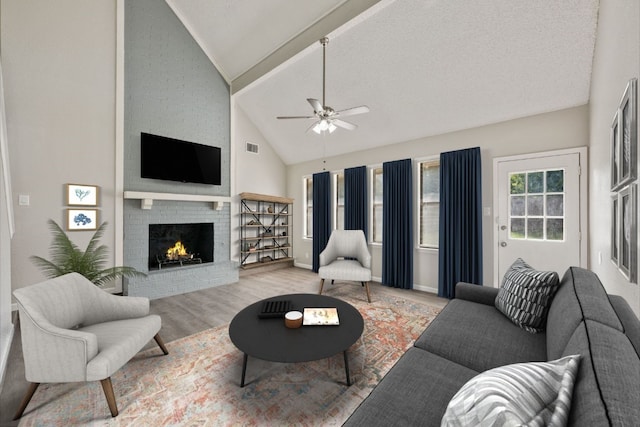 living room with a textured ceiling, ceiling fan, high vaulted ceiling, light hardwood / wood-style floors, and a brick fireplace