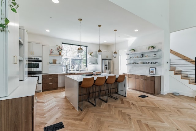 kitchen featuring appliances with stainless steel finishes, brown cabinets, a kitchen island with sink, light countertops, and open shelves