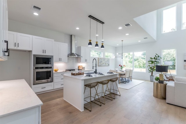 kitchen featuring wall chimney range hood, a kitchen island with sink, white cabinets, appliances with stainless steel finishes, and light hardwood / wood-style floors