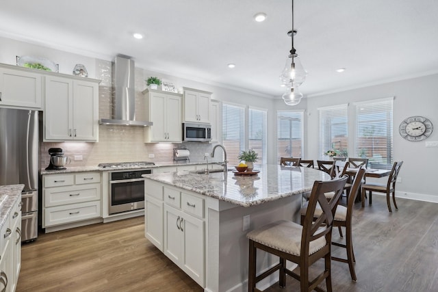 kitchen with appliances with stainless steel finishes, a kitchen island with sink, wall chimney range hood, sink, and wood-type flooring