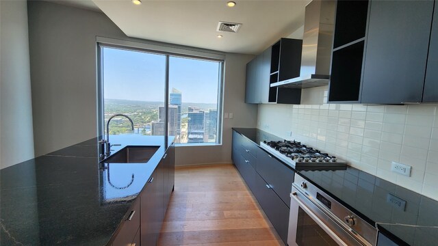 kitchen with light hardwood / wood-style flooring, wall chimney range hood, dark stone countertops, backsplash, and sink