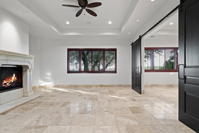 unfurnished living room with ceiling fan, a premium fireplace, a barn door, and a raised ceiling