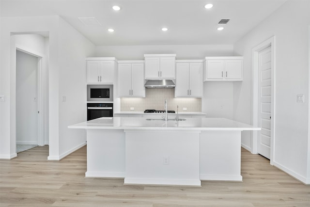 kitchen with white cabinetry, tasteful backsplash, a center island with sink, built in microwave, and oven