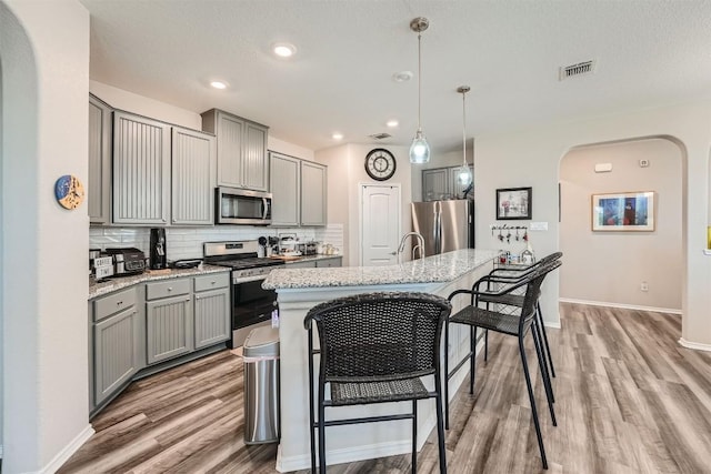 kitchen with pendant lighting, gray cabinetry, backsplash, a kitchen island with sink, and appliances with stainless steel finishes