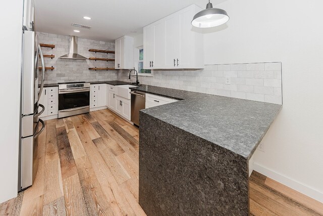 kitchen with appliances with stainless steel finishes, tasteful backsplash, kitchen peninsula, wall chimney range hood, and light wood-type flooring