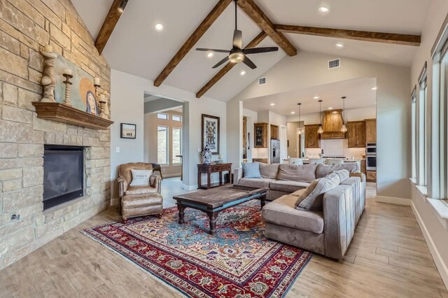 living room featuring ceiling fan, light wood-type flooring, a stone fireplace, high vaulted ceiling, and beam ceiling