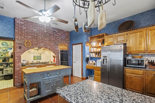 kitchen with ceiling fan, brick wall, appliances with stainless steel finishes, a center island, and dark tile patterned floors