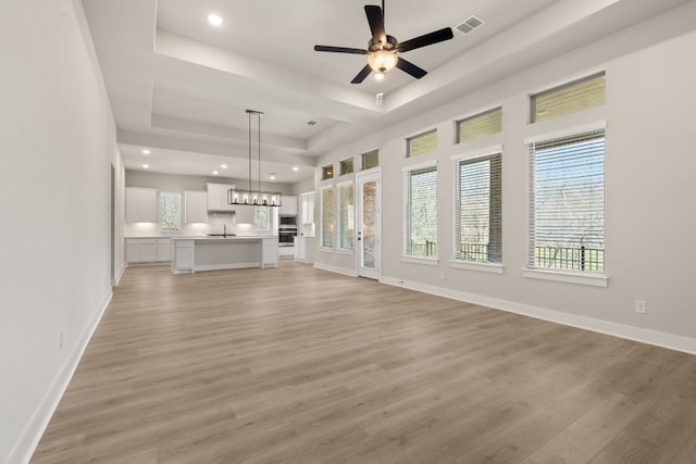 unfurnished living room featuring ceiling fan with notable chandelier, light wood-type flooring, a raised ceiling, and baseboards