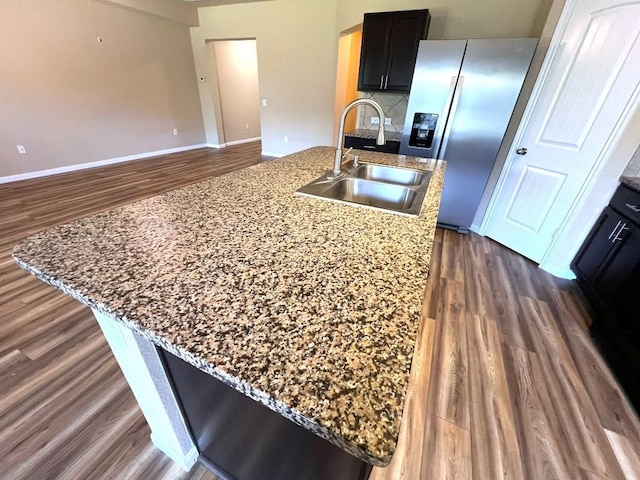 kitchen featuring light stone counters, dark wood-type flooring, a sink, an island with sink, and stainless steel fridge