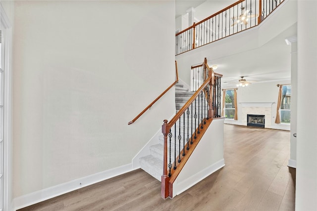 stairway featuring wood finished floors, a towering ceiling, a fireplace, baseboards, and ceiling fan