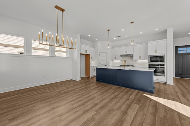 kitchen with stainless steel oven, built in microwave, decorative light fixtures, a center island with sink, and white cabinets