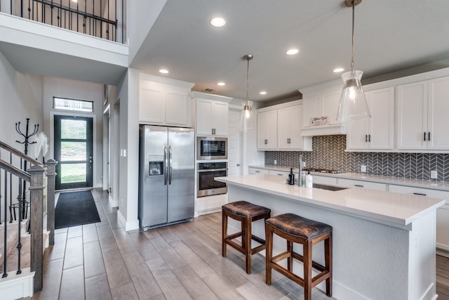 kitchen with a kitchen island with sink, stainless steel appliances, hanging light fixtures, white cabinetry, and light hardwood / wood-style flooring