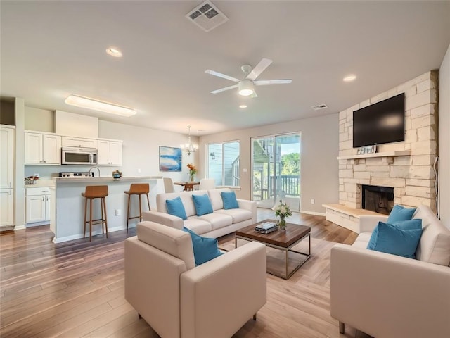 living room with ceiling fan with notable chandelier, a stone fireplace, and light wood-type flooring