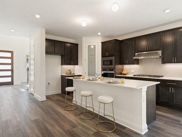 kitchen featuring decorative backsplash, dark wood-type flooring, an island with sink, and stainless steel appliances