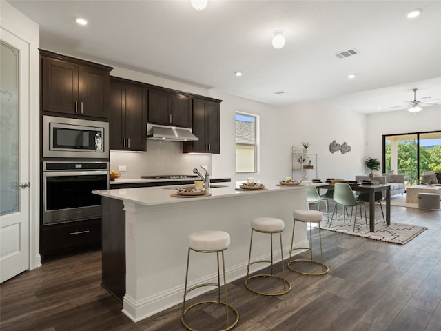 kitchen with ceiling fan, decorative backsplash, dark hardwood / wood-style floors, an island with sink, and stainless steel appliances