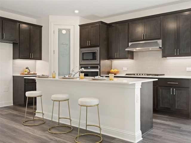 kitchen with dark wood-type flooring, dark brown cabinets, tasteful backsplash, stainless steel appliances, and a center island with sink