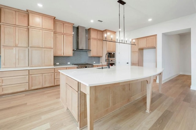 kitchen with wall chimney exhaust hood, hanging light fixtures, an island with sink, light hardwood / wood-style floors, and light brown cabinetry