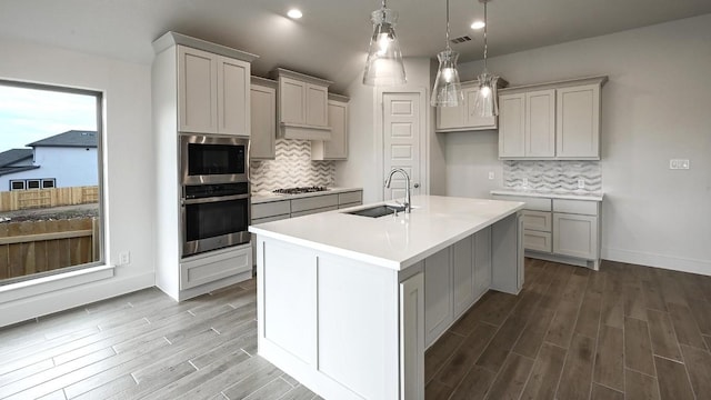 kitchen with decorative backsplash, appliances with stainless steel finishes, a kitchen island with sink, sink, and hanging light fixtures